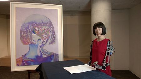 Ai-Da, the artificially intelligent robot, poses next to her self-portrait currently on display in the London Design Museum.