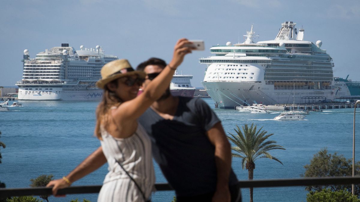 زوجان يلتقطان صورة سيلفي في ميناء مدينة ميورقة الإسبانية وخلفهما سفنٌ سياحية راسية