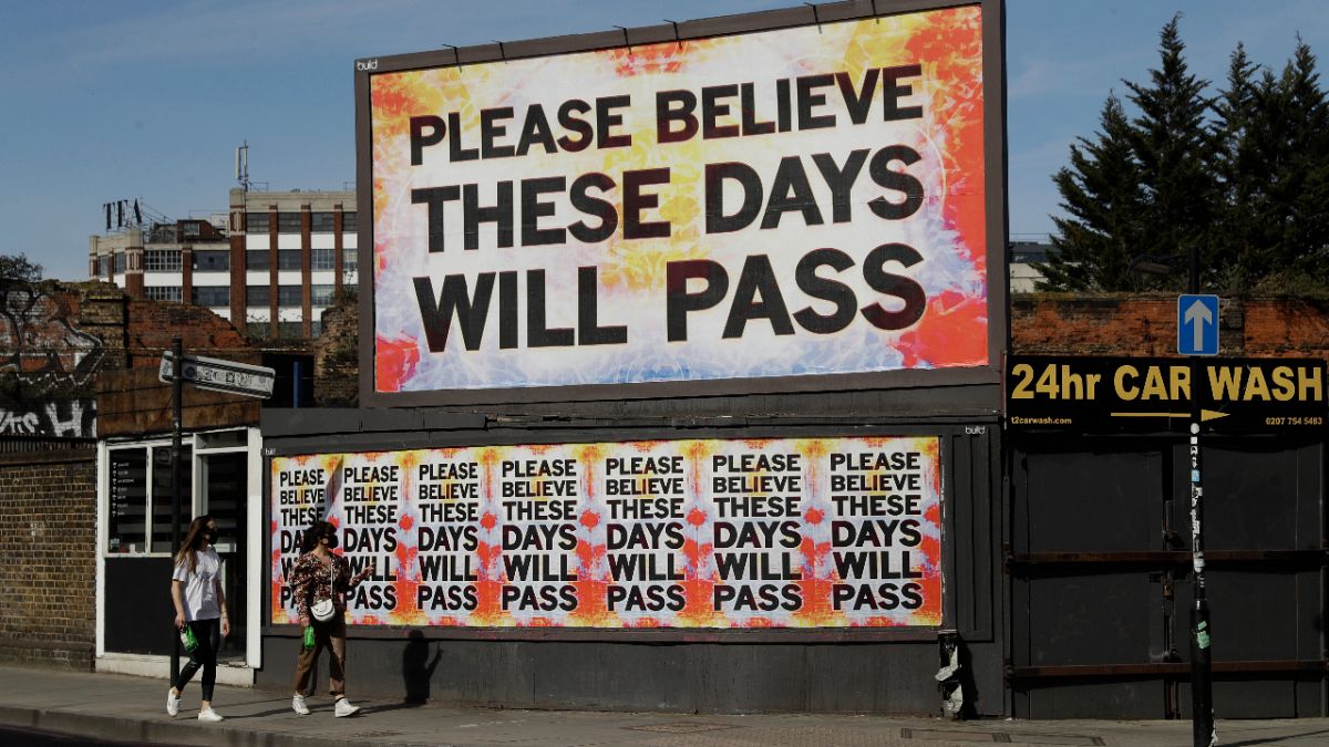 Women wearing face masks to protect from coronavirus walk past a billboard with a work by artist Mark Titchner in east London, Tuesday, April 7, 2020. 