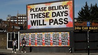 Women wearing face masks to protect from coronavirus walk past a billboard with a work by artist Mark Titchner in east London, Tuesday, April 7, 2020. 