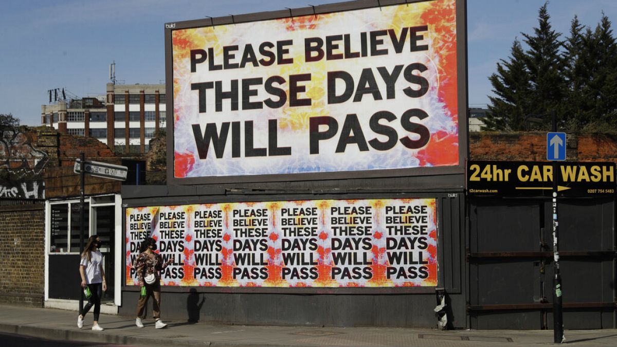 Mujeres con mascarillas para protegerse del coronavirus pasan junto a una valla publicitaria con una obra del artista Mark Titchner en el este de Londres