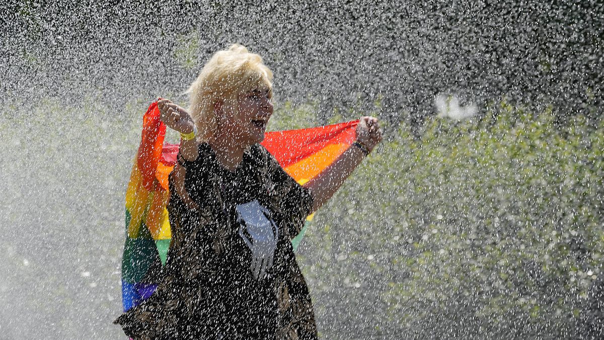 A woman with a rainbow flag cools off in a sprinkler ahead of the Equality Parade, the largest LGBT pride parade. Warsaw, Poland. June 19, 2021