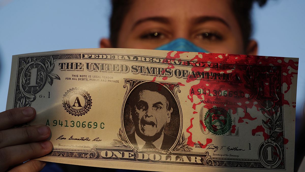 Manifestante segura cópia de dólar americano sobreposto com a imagem do Presidente do Brasil, marcada com tinta vermelha para simbolizar o sangue das vítimas de Covid-19