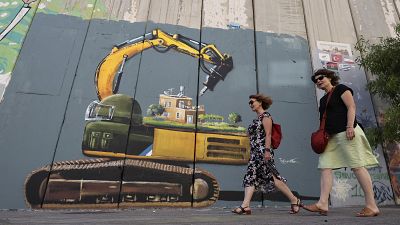 سائحون يمرون بجانب لوحة جدارية تصور جنديًا إسرائيليًا مع جرافة على وشك هدم منزل فلسطيني، رسمها الفنان الفلسطيني تقي سباتين