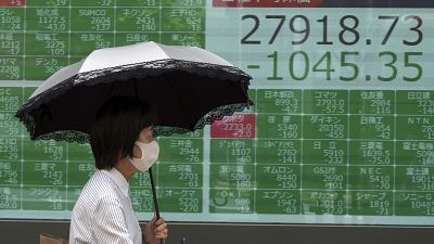 Ernyőt tart a feje fölé egy tőzsdei árfolyamokat mutató tábla előtt elsétáló férfi Tokióban 2021. június 21-én