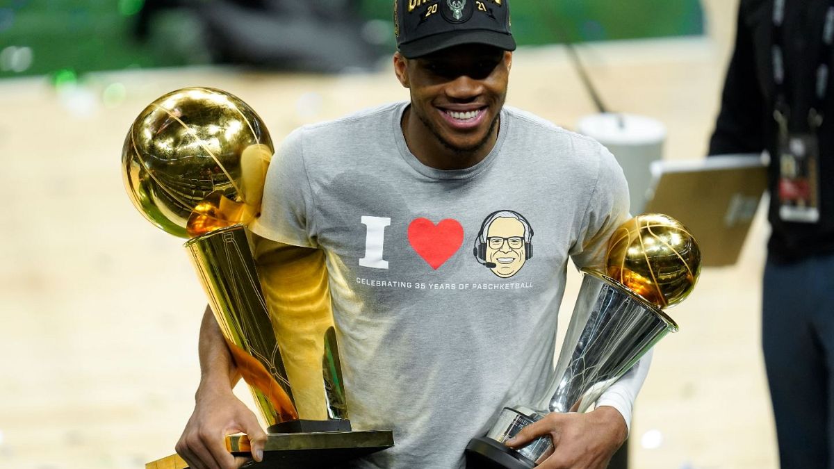Giannis Antetokounmpo smiles while holding the NBA Championship trophy and Most Valuable Player trophy.