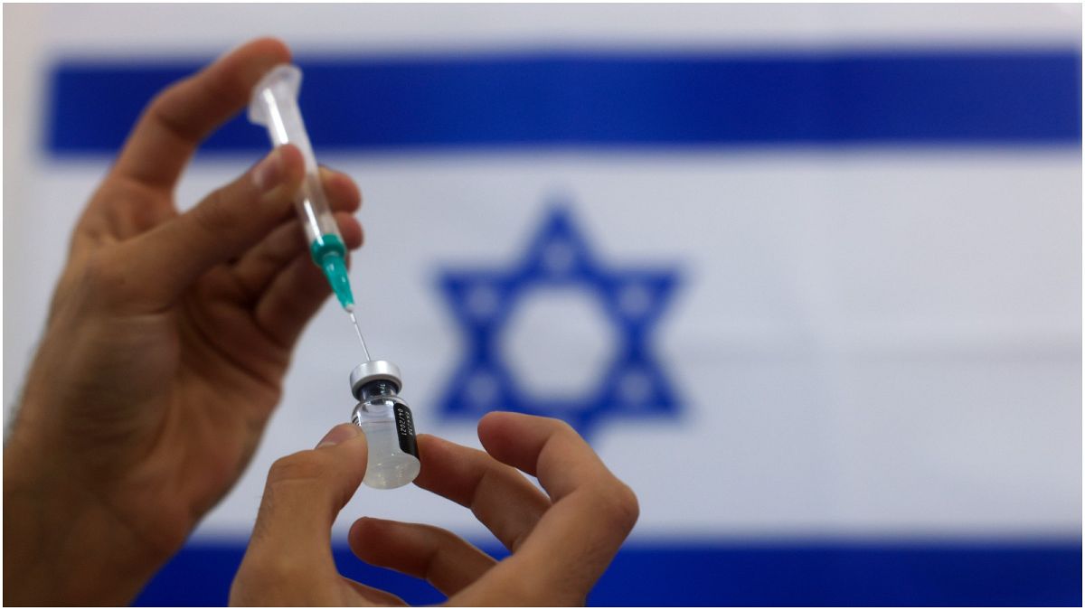 موظفٌ في القطاع الصحي الإسرائيلي يحضّر جرعة لقاح ضد كورونا  وفي الخلفية علم إسرائيل