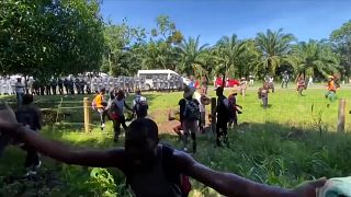 Shielded guardsmen facing migrants, one throwing a rock