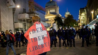 A man holding a Swiss flag on his shoulders reading "Freedom" walks past riot police.