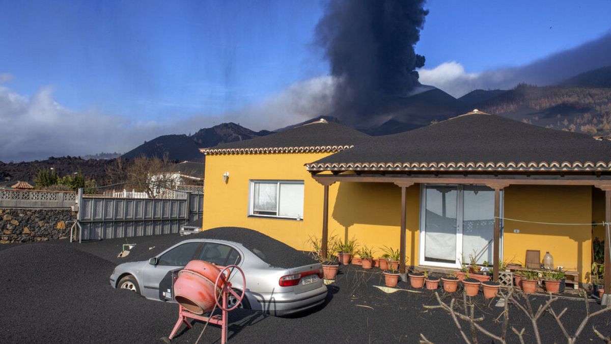 Imagen de una vivienda parcialmente cubierta con ceniza volcánica y el volcán Cumbre Vieja al fondo, en la isla de La Palma.