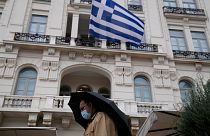 A man wearing a face mask to protect against coronavirus, walks outside a store as a Greek flag waves in Athens, Greece.