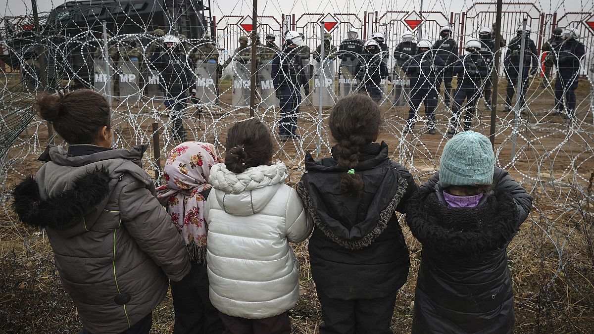 Migrants children stand in front of a barbed wire fence and Polish servicemen at the checkpoint "Kuznitsa" at the Belarus-Poland border, Belarus, Nov. 17, 2021.