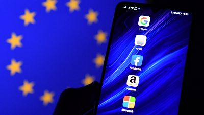 Logos of Google, Apple, Facebook, Amazon and Microsoft displayed on a mobile phone with an EU flag displayed in the background