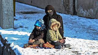 سيدة تجلس مع طفليها على جانب طريق مغطى بالثلوج في كابول