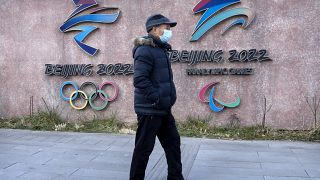 A man wearing a face mask walks past the logos for the Beijing 2022 Winter Olympics and Paralympics in Beijing, Thursday, Dec. 16, 2021.