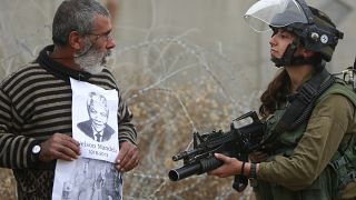 متظاهر فلسطيني يحمل صور الزعيم الجنوب أفريقي الراحل نيلسون مانديلا والزعيم الفلسطيني الراحل ياسر عرفات أمام جندي إسرائيلي. 2013/12/06