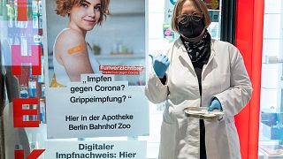 A pharmacist stands outside the pharmacy with vaccination paraphernalia, next to a poster advertising the Corona vaccination in Berlin, Germany, Feb. 7, 2022.