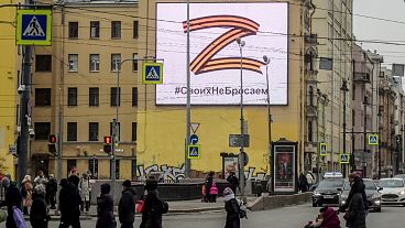 Pedestrians cross a street in front of a billboard displaying the symbol Z in the colours of the ribbon of Saint George and a slogan reading: "We don't give up on our people".