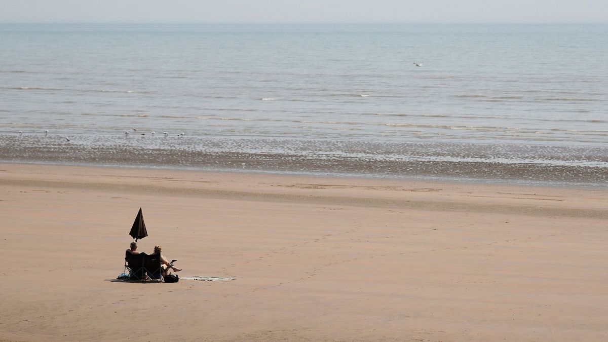 Two sunbathers sit and watch the sea on the sandy beach at Camber Sands, England.