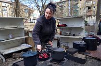 Γυναίκα μαγειρεύει σε αυτοσχέδιο κουζίνα στο ύπαιθρο με φόντο τα κατεστραμμένα κτίρια της Μαριούπολης