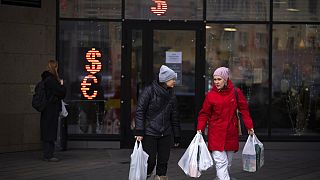 Two women walk past a currency exchange office screen displaying the exchange rates of U.S. Dollar and Euro to Russian Rubles in Moscow
