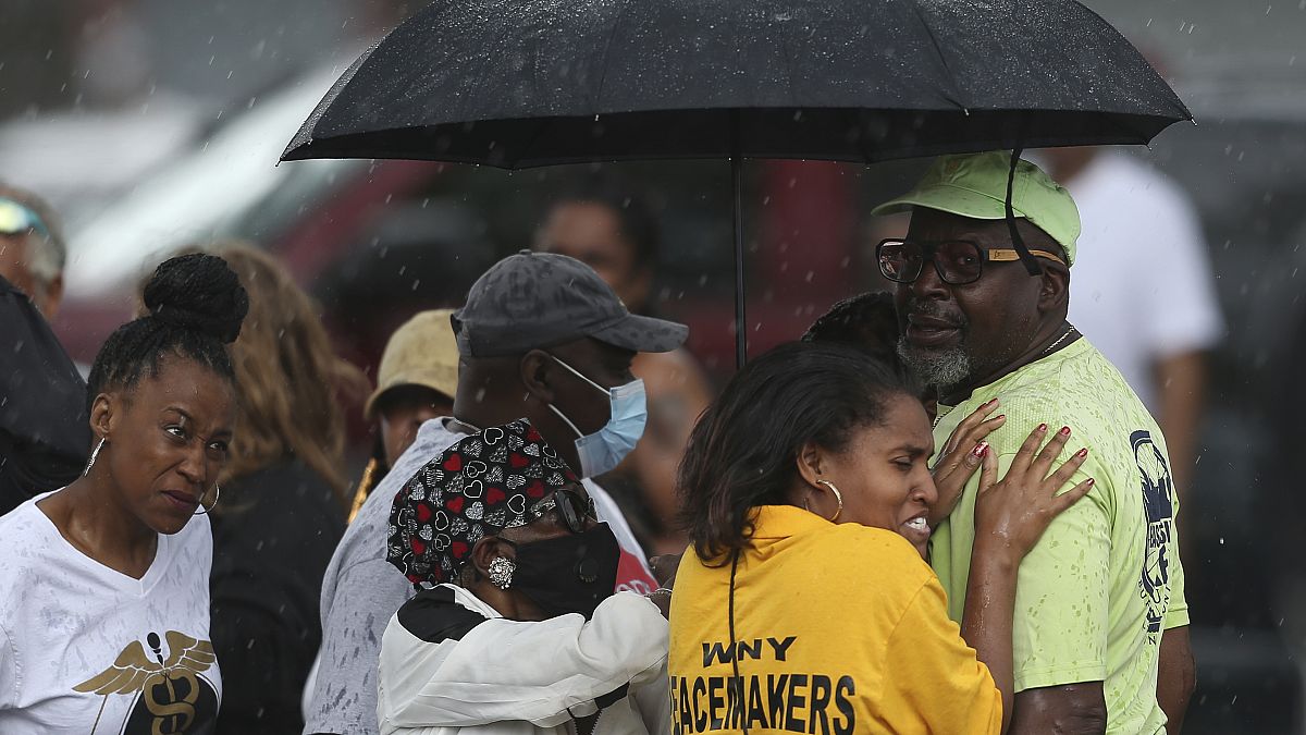 Bystanders gather under an umbrella as rain rolls in after a shooting at a supermarket on Saturday, May 14, 2022, in Buffalo, N.Y.