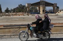 راكبان على دراجة نارية مقابل مدرسة سابقة كانت تابعة لتنظيم الدولة الإسلامية في الرقة