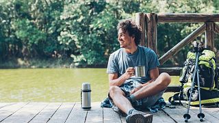 A traveller sits in by a lake with his reusable coffee cup