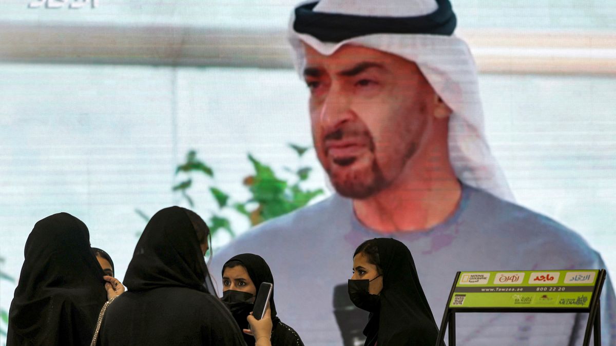 سيدات يتصفحن صحيفة قبالة صورة لرئيس الإمارات الجديد الشيخ محمد بن زايد آل نهيان في معرض أبوظبي الدولي للكتاب 2022.