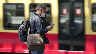 Egy utas tájékozódik mobiltelefonjából, miközben egy elővárosi vonat érkezik a pályaudvarra