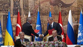 El presidente turco y el secretario general de la ONU se sientan mientras dos representantes de las delegaciones de Ucrania y Rusia se dan la mano