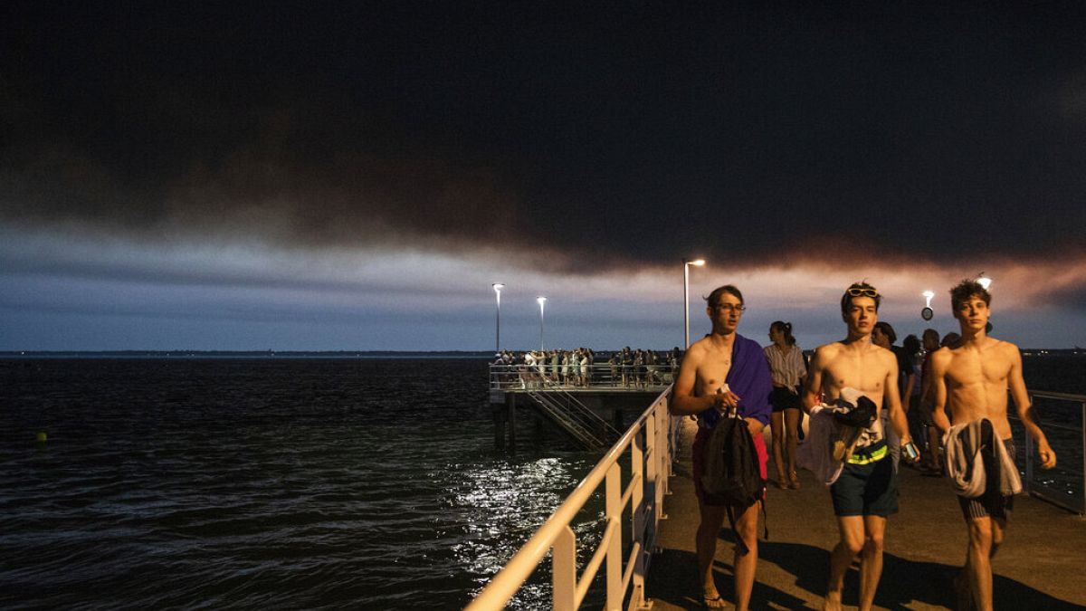 Swimmers walk on the Le Moulleau pier in Arcachon, southwestern France Monday July 18, 2022, as a large cloud of black smoke laden with ashes coming from a giant wildfire.