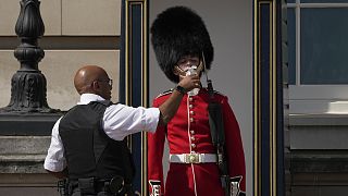 A police officer givers water to a British soldier wearing a traditional bearskin hat, on guard duty outside Buckingham Palace on 18 July 2022
