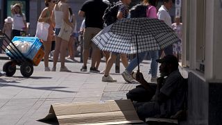 Un hombre se sienta bajo un paraguas para resguardarse del sol mientras pide dinero en la calle durante la ola de calor en Madrid, España, el sábado 16 de julio de 2022