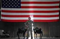 جندي أمريكي يقف بين كلبين آليين وخلفه علم الولايات المتحدة، 11 أغسطس 2022.