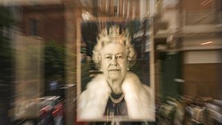 Emberek sétálnak el II. Erzsébet királynő portréja mellett egy London központjában lévő üzletben