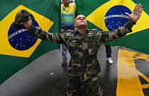 A supporter of President Jair Bolsonaro dressed in fatigues, kneels with his arms spread out in front of Brazilian national flags