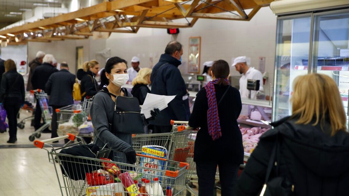 A woman wearing a face mask holds a trolley as other customers wait in a queue to shop goods at a supermarket in Athens