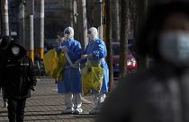 People walk by workers in protective suits look for the location of the lockdown residents to collect COVID samples in Beijing, 1 December 2022