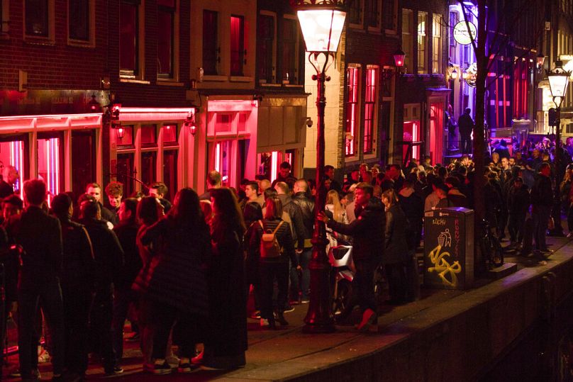 In this Friday March 29, 2019, file image tourists bathing in a red glow emanating from the windows and peep shows' neon lights, in Amsterdam's red light district.