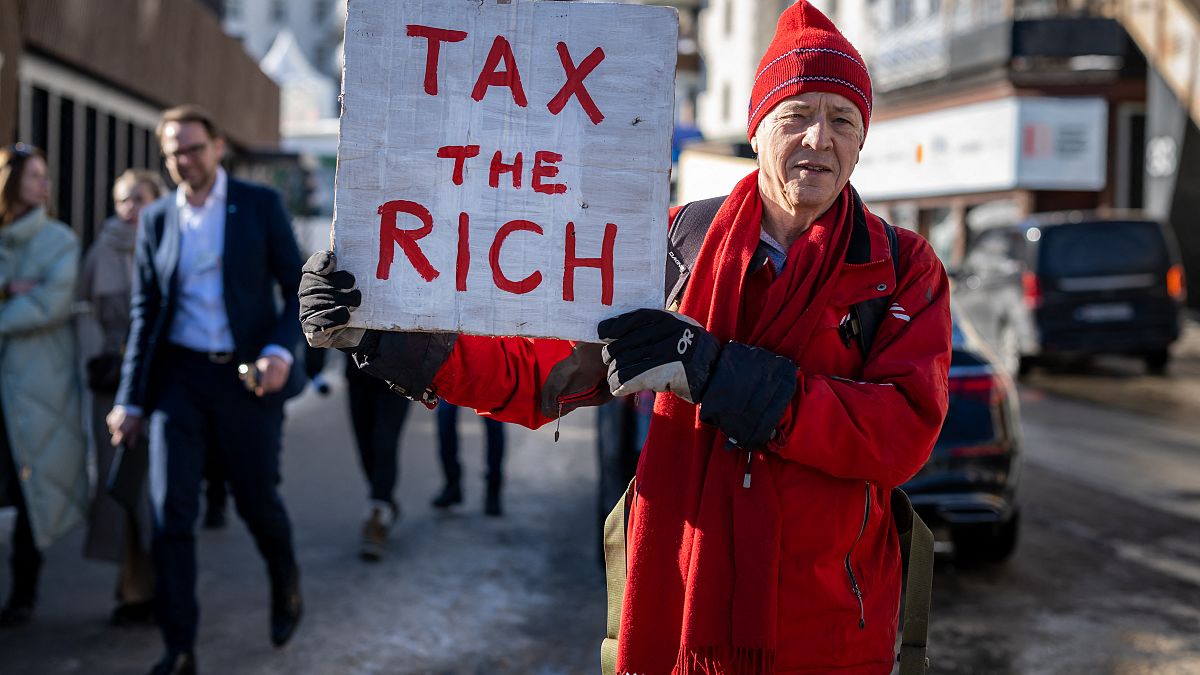 هيل وايت، مليونير بريطاني يقف مع لافتة كتب عليها: "إفرضوا ضرائب على الأغنياء" بجوار مقر الاجتماع السنوي لمنتدى في دافوس في سويسرا، في 18 يناير/كامون الثاني 2023. 