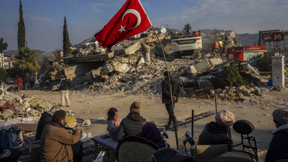 Família senta-se ao lado de um edifício desmoronado enquanto espera que os corpos de familiares sejam recuperados dos escombros em Antakya, Turquia
