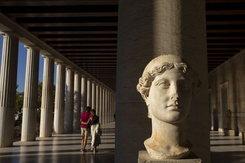 A young couple walk behind a 2nd century A.D. marble head of victory inside Stoa of Attalos, at ancient Agora, in Athens, October 2022