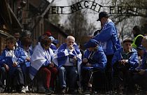 Holokauszt-túlélők ülnek a hírhedt auschwitzi felirat alatt
