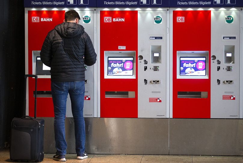 A man buys a train ticket as writing reads "All you can drive! - 49 Euro the Germany Ticket" on the monitors of ticket vending machines at the main train station in Cologne.