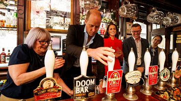Vilmos herceg sört csapol egy londoni pubban, mögötte piros ruhában Katalin hercegnő 2023.05.04-én. 