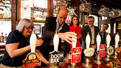Vilmos herceg sört csapol egy londoni pubban, mögötte piros ruhában Katalin hercegnő 2023.05.04-én. 
