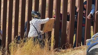 Un repartidor entregando una pìzza desde México a través de los barrotes de la valla fronteriza con EEUU