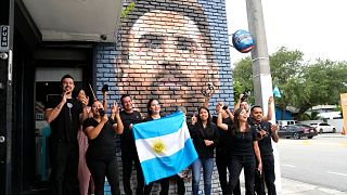 Staff at the Fiorito restaurant hold an Argentinian flag as they pose for a photograph taken by a colleague in front of a mural of Lionel Messi, Wednesday, June 7, 2023, in Mi