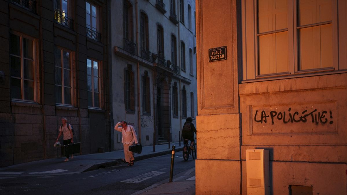 People pass by a wall illuminated by a burning barricade on which is written 'Police kills' in the center of Lyon, central France, Friday, June 30, 2023. 